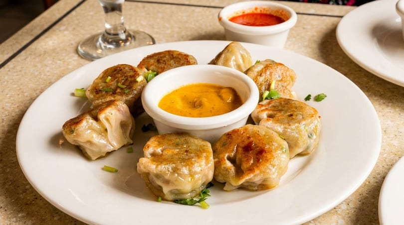 A white plate holds pan-fried dumplings with two dipping sauces, one yellow and one red.