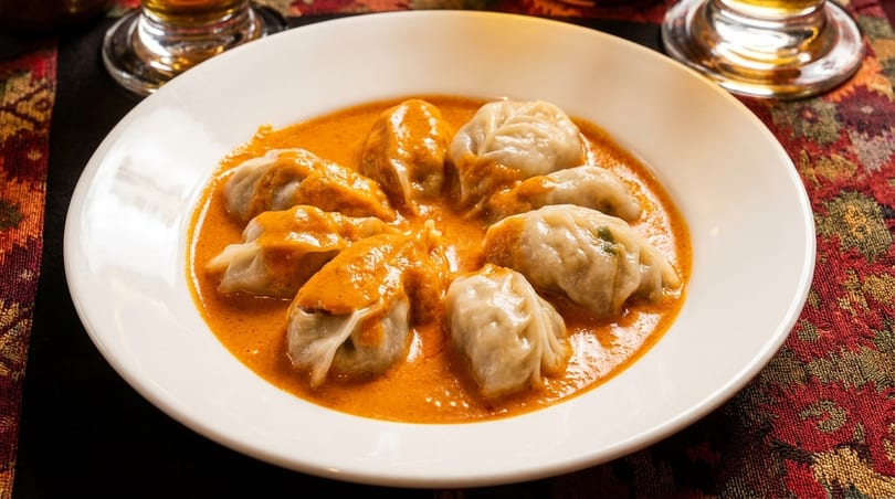 A white bowl filled with momos in a rich orange sauce, on a patterned tablecloth.